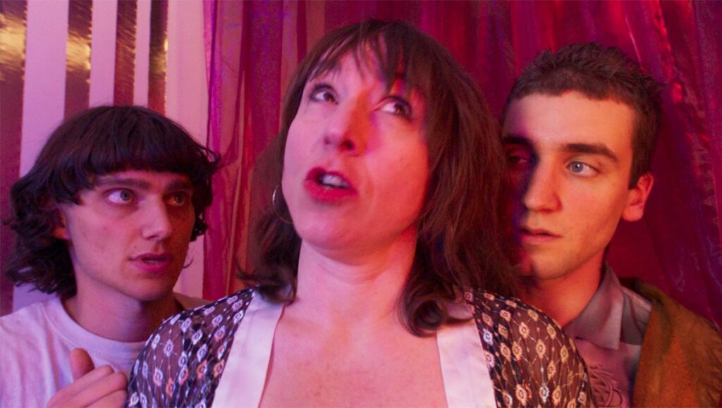 Mother Schmuckers — Harpo & Lenny Guit Prime Time film still. Woman between two men, lit in pink and red.