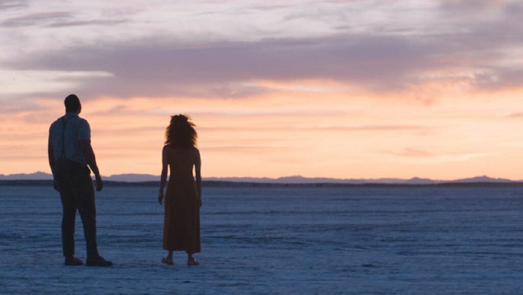 Nine Days | Edson Oda Nine Days film still. Two figures silhouetted against a sunset sky over a vast, desolate landscape. Edson Oda movie.