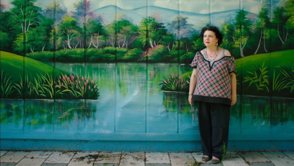 Some Kind of Heaven film still: Woman stands before a mural of a lake and trees.
