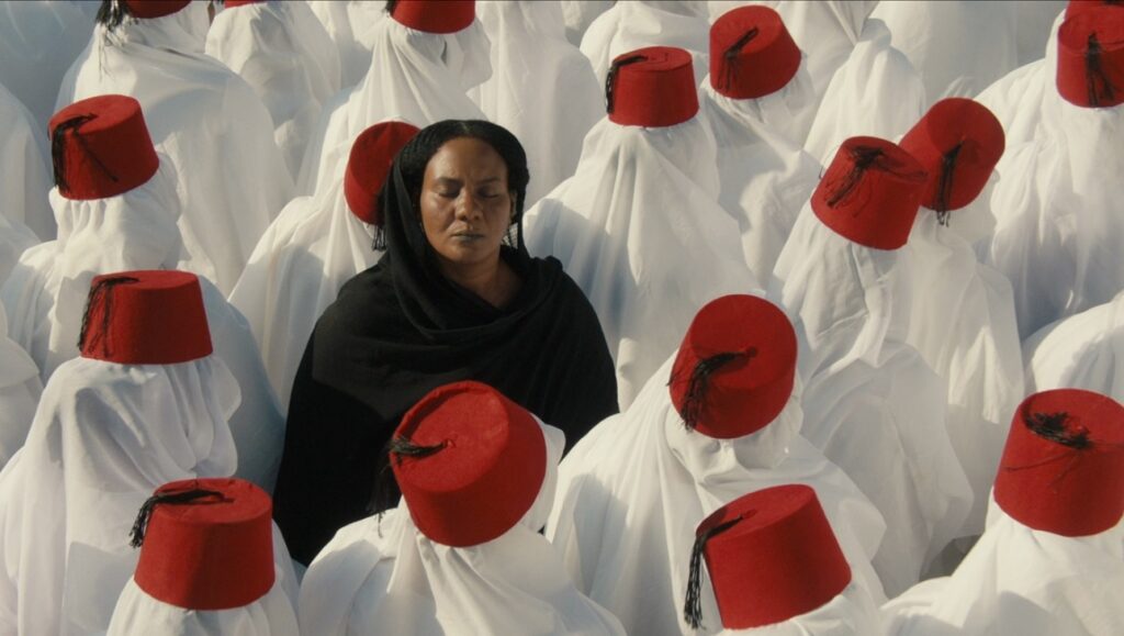 You Will Die at Twenty film still. Woman in black surrounded by figures in white robes and red fez hats.