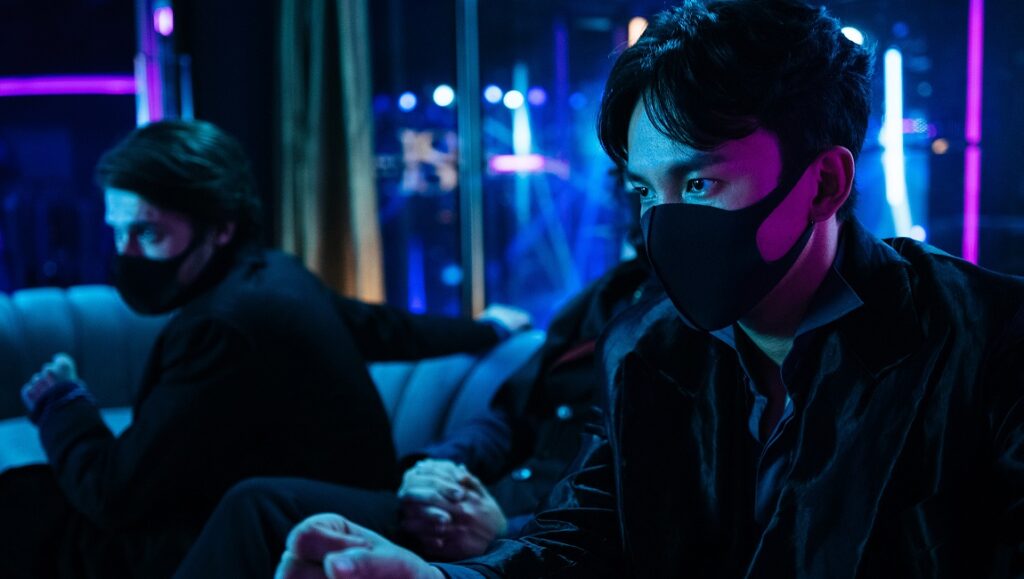 Dead & Beautiful | David Verbeek Dead & Beautiful film still. Two masked men in dark suits sit on a couch in a room with blue and purple lighting. IFFR 2021.