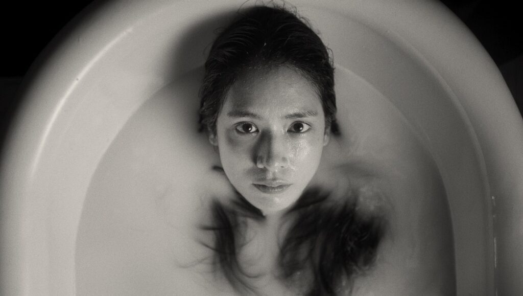Film still from The Edge of Daybreak: Woman in a bathtub, looking up. Black and white. Taiki Sakpisit.
