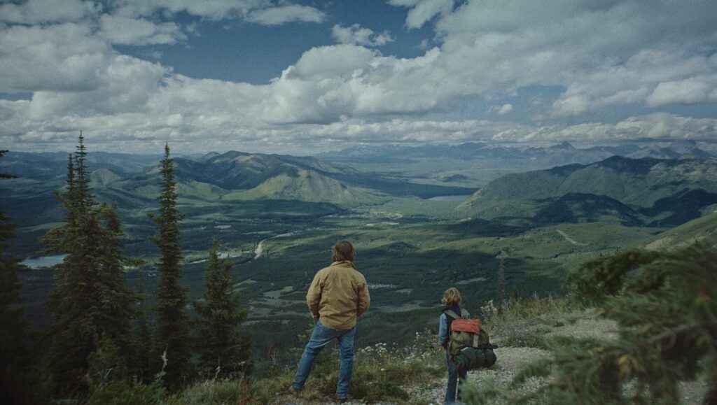 Cowboys movie scene: Two people overlook a vast mountain range landscape under a cloudy sky.