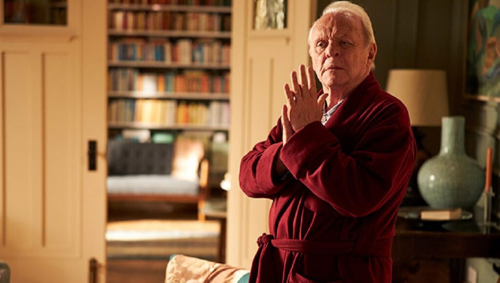 Anthony Hopkins in The Father movie. Hopkins wears a red robe, hands clasped, in a room with a bookshelf.
