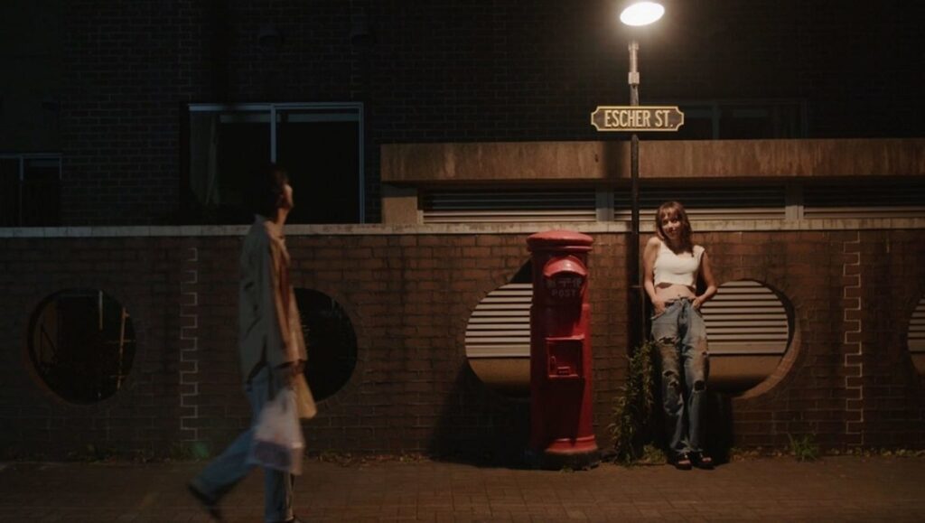 Red Post on Escher Street | Sion Sono Red Post on Escher Street film still. Woman by red postbox under street sign. Sion Sono movie scene.