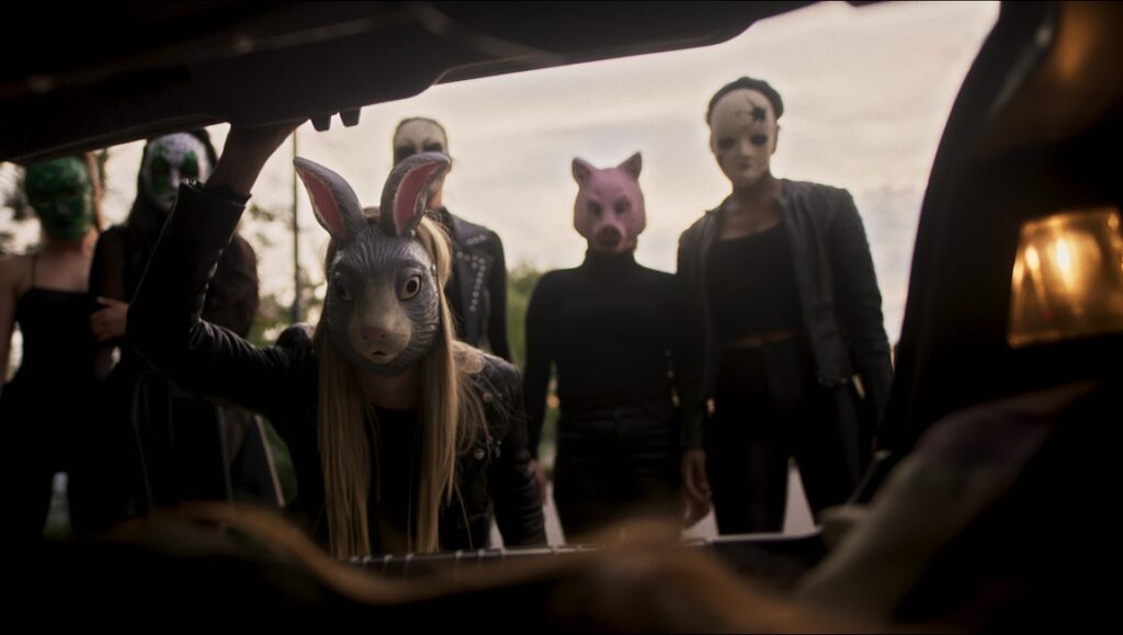 The Sinners movie scene: People in animal masks, including a rabbit mask, stand behind an open car trunk.