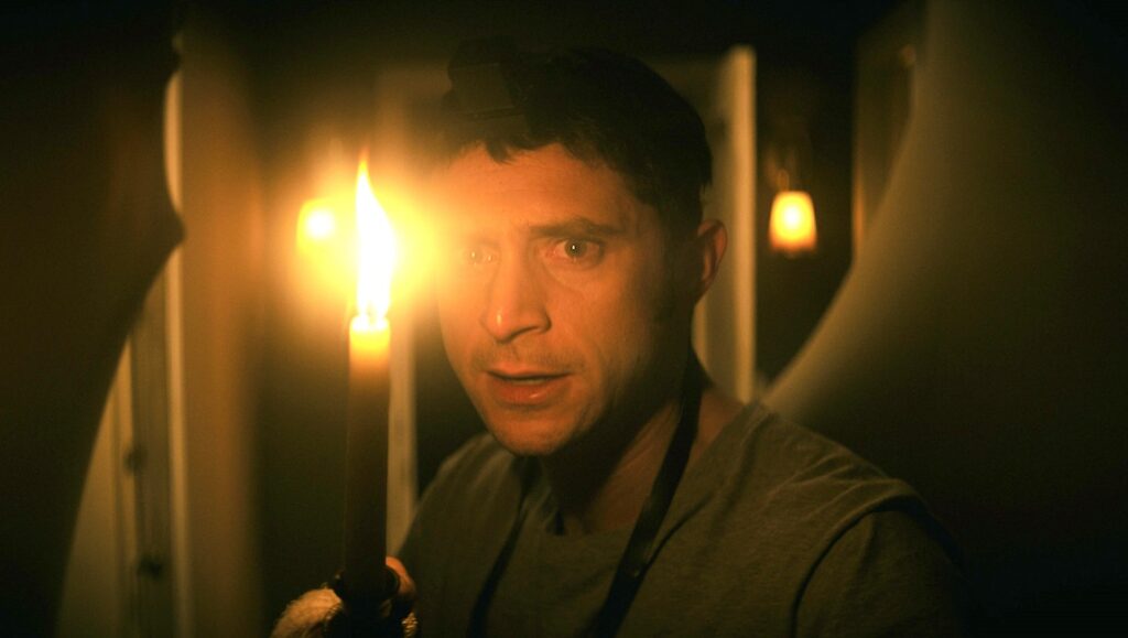 Still from The Vigil, a horror film. Man holding candle, looking scared in a dimly lit room.