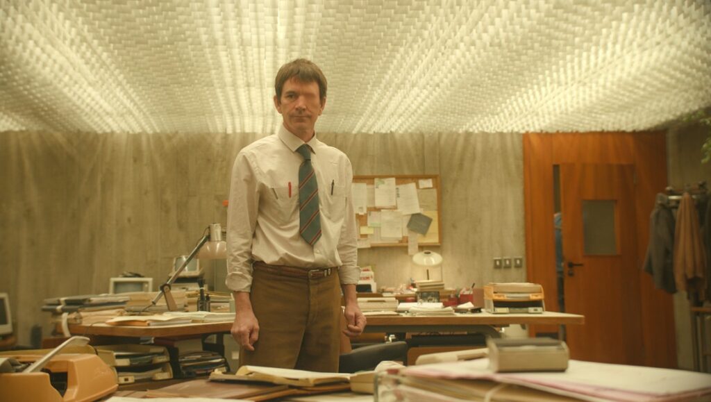 Keep an Eye Out | Quentin Dupieux Keep an Eye Out film still: Man in office. Quentin Dupieux movie scene with a cluttered desk and unique ceiling.