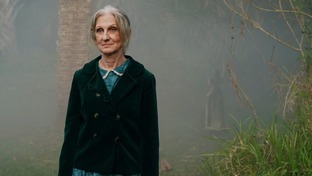Offseason — Mickey Keating Still from Offseason: An older woman in a green jacket stands in a misty, overgrown area.