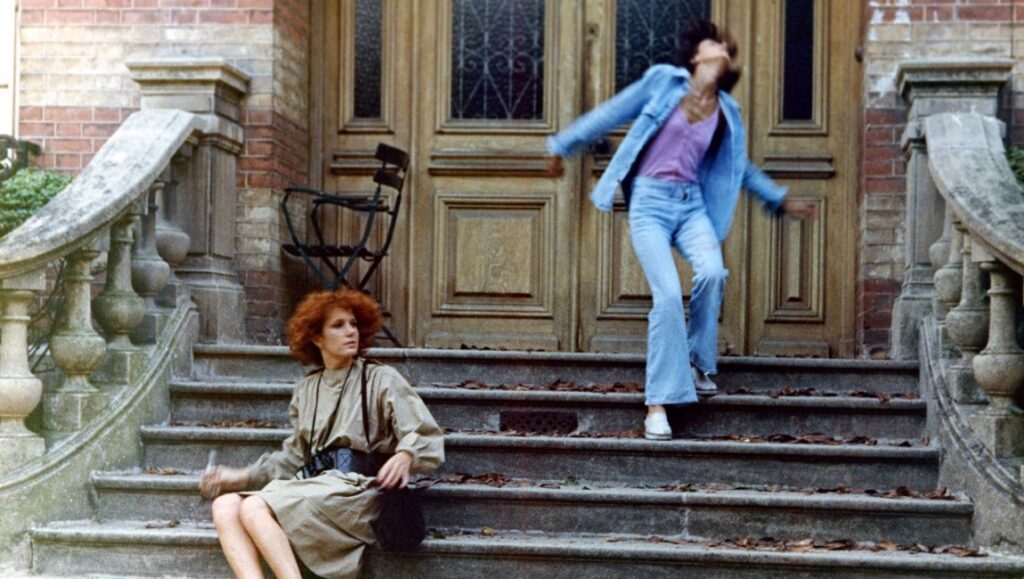 Celine and Julie Go Boating | Jacques Rivette Celine and Julie Go Boating film still. Two women on stone steps in front of a large wooden door. Jacques Rivette film.