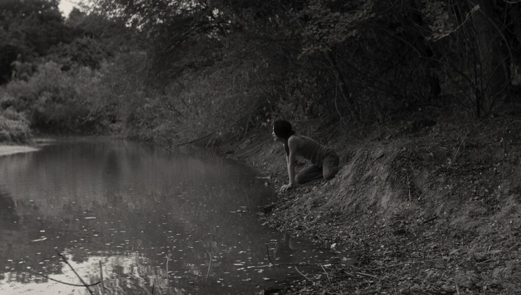 Come Here | Anocha Suwichakornpong Still from Come Here at Berlin Film Festival. Woman kneels at river's edge. Black and white.