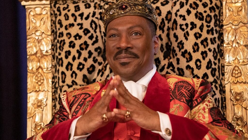 Coming 2 America | Craig Brewer Eddie Murphy as King Akeem in Coming 2 America. Regal, wearing a crown and red suit. Seated on a leopard print throne.