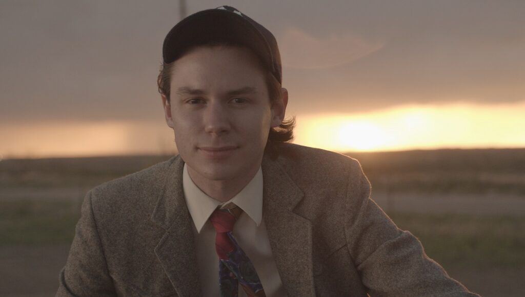 Kid Candidate film still from SXSW 2021. Young man in suit and baseball cap smiles against a sunset backdrop.
