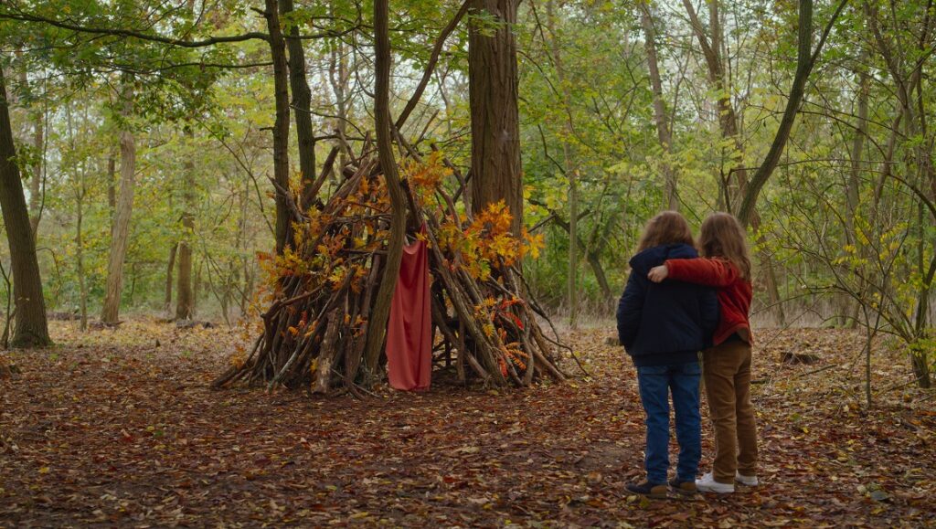 Petite Maman — Celine Sciamma Petite Maman film still: Two young girls embrace near a stick fort in a forest. Berlin Film Festival 2021.