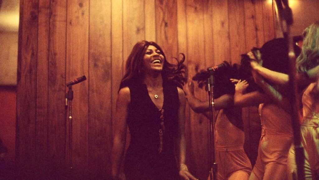 Tina Turner performing live. The singer is smiling and wearing a black dress. Microphones and dancers are visible.