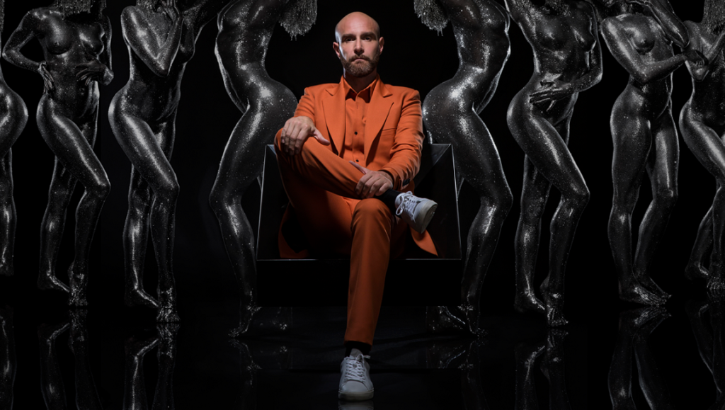 Vitalic, OK Cowboy album art. Musician in orange suit surrounded by dark, glossy figures. Electronic music artist promo.