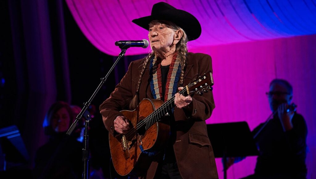 Willie Nelson performs live in February 2021. The country music star plays guitar on stage in a black cowboy hat.