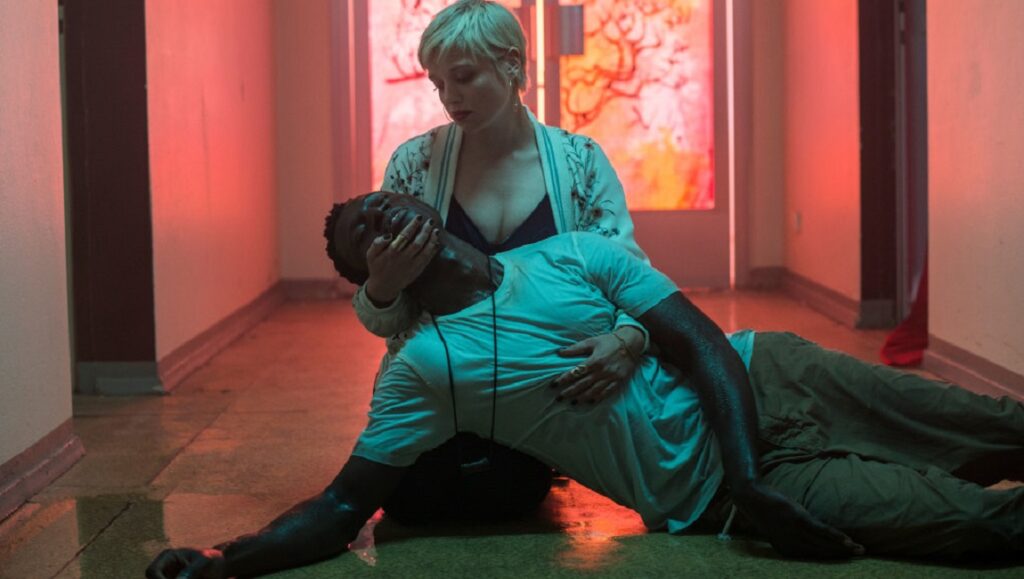 Berlin Alexanderplatz | Burhan Qurbani Things Heard and Seen movie scene. Woman holds man lying on floor. Hallway with red and green lighting.