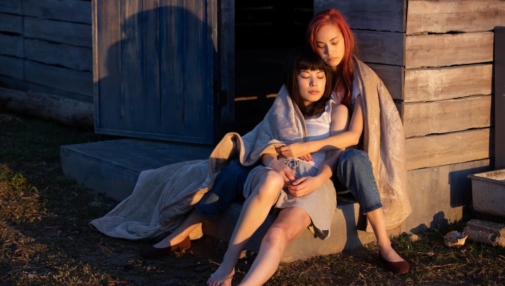 Ride or Die film: Two women embrace, wrapped in a blanket outside a rustic building. Ryuichi Hiroki directs this drama.