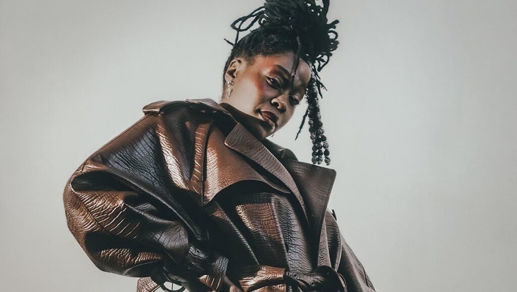 BbyMutha | Muthaleficent 2 BBymutha album review. Portrait of BBymutha in a brown leather trench coat, hair in a high bun of dreadlocks.