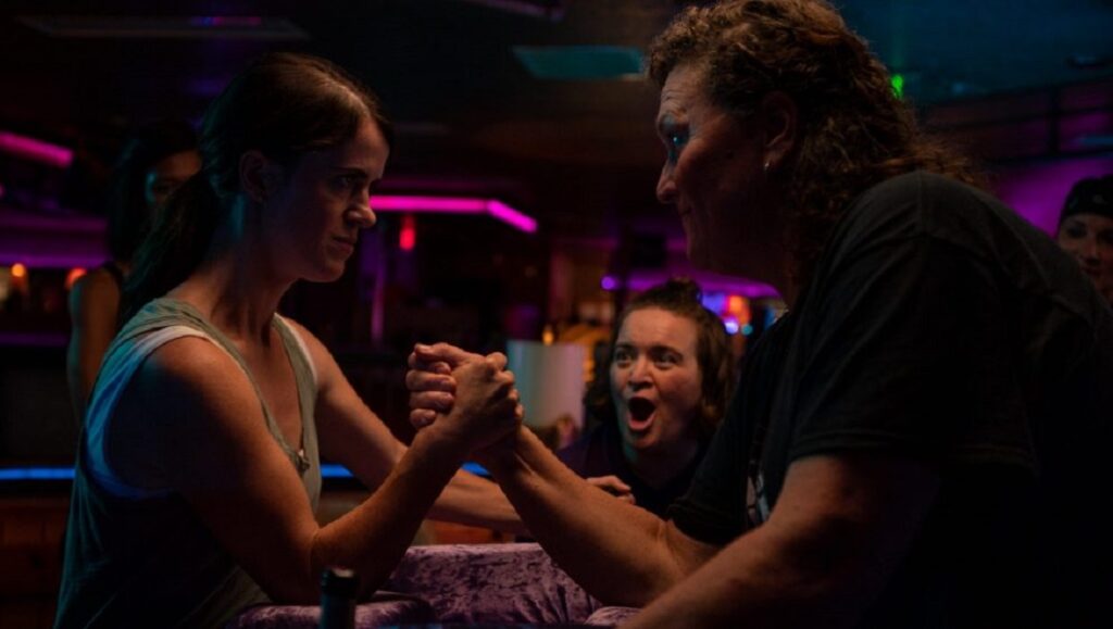 Golden Arm | Maureen Bharoocha Golden Arm movie scene: Two women arm wrestling, intense competition. Crowd watches.