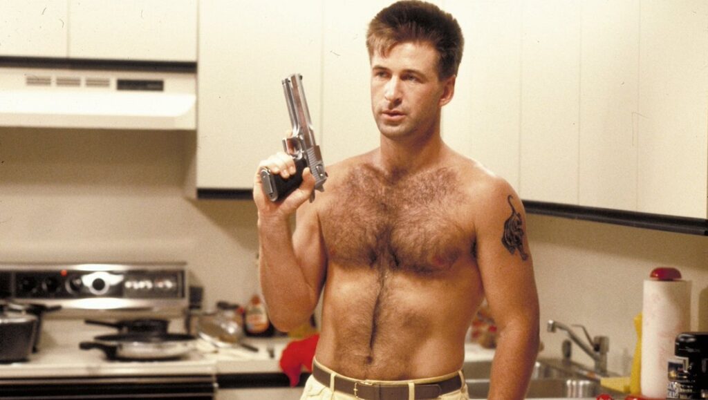 Alec Baldwin in Miami Blues movie review. Shirtless actor holding a gun in a kitchen.