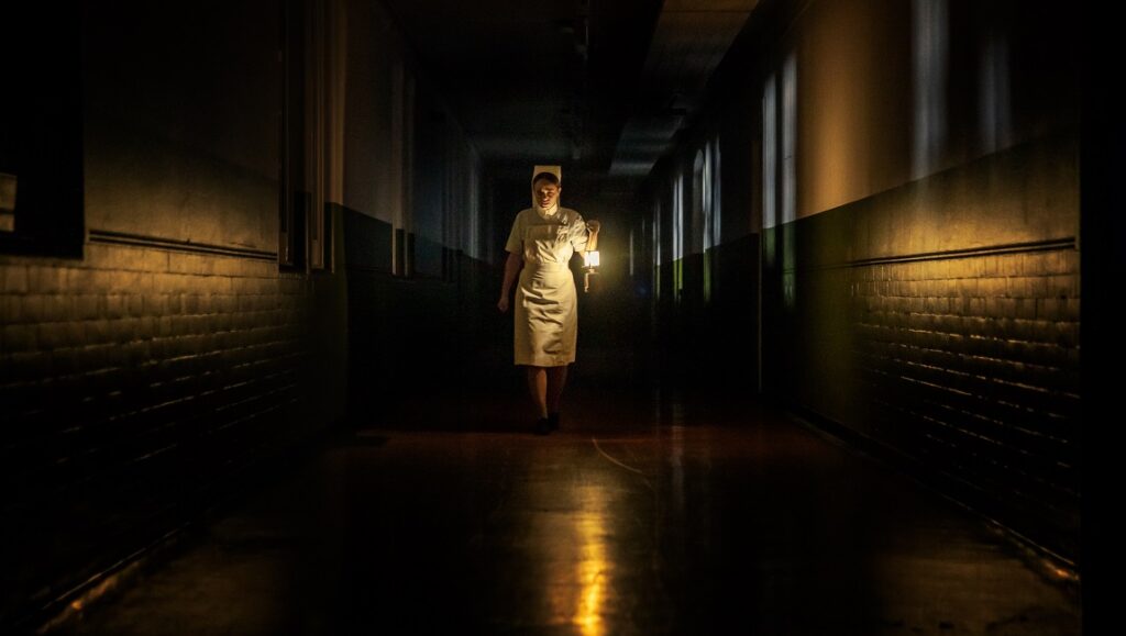 The Power | Corinna Faith The Power film still: Nurse in a dark hospital hallway holding a lantern. Horror movie scene.