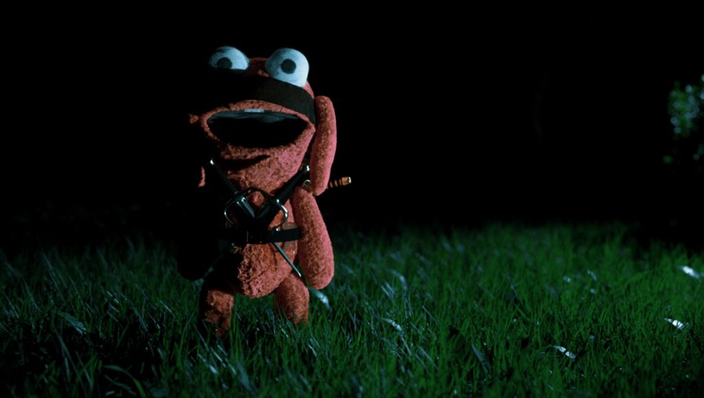 Benny Loves You | Karl Holt Stuffed animal ninja. Red plush toy with a sword, blindfold, and harness stands in grass at night. Before We Vanish movie.