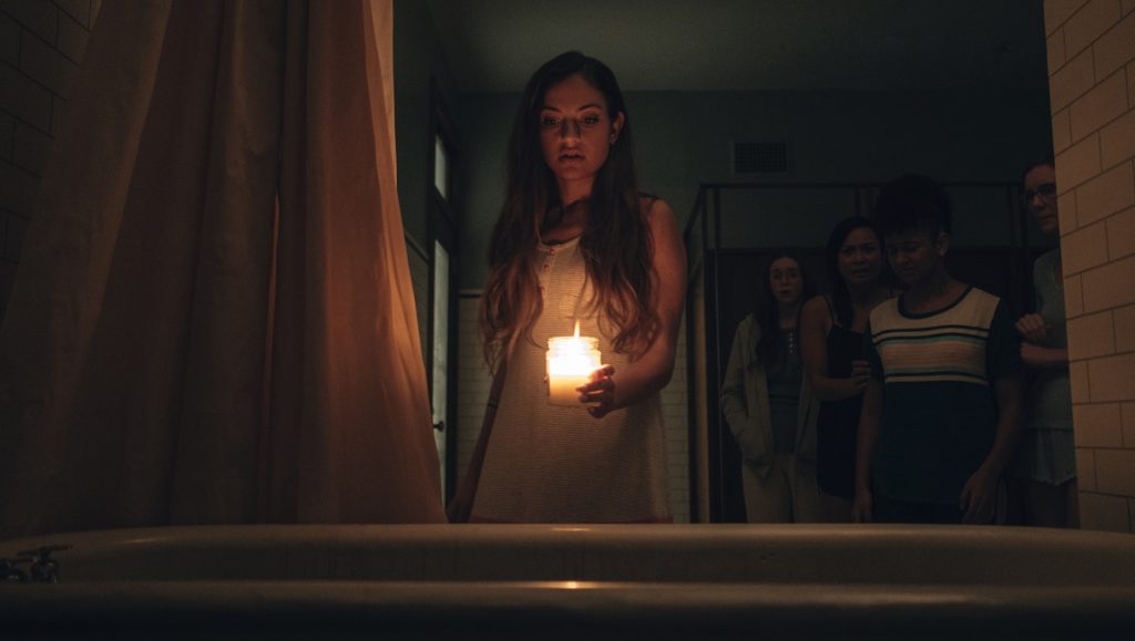 Seance | Simon Barrett Seance movie still. Woman holding candle in dark bathroom, other students behind her. Horror film scene.