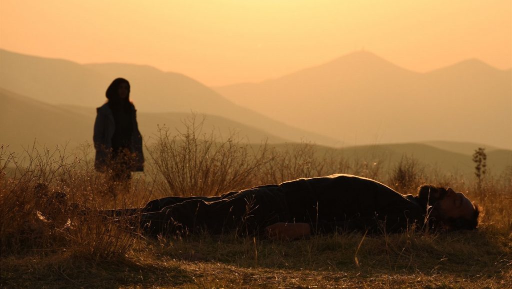 There is No Evil film still. Silhouette of person standing over body in field, mountains in background.