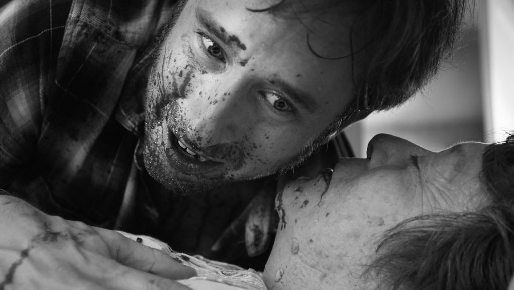 Dementia Part II film still. A man with a dirty face looms over an older person lying down. Black and white.