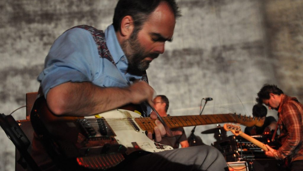 Godspeed You! Black Emperor performing live. Band member playing electric guitar on stage. April 2021 album roundup.