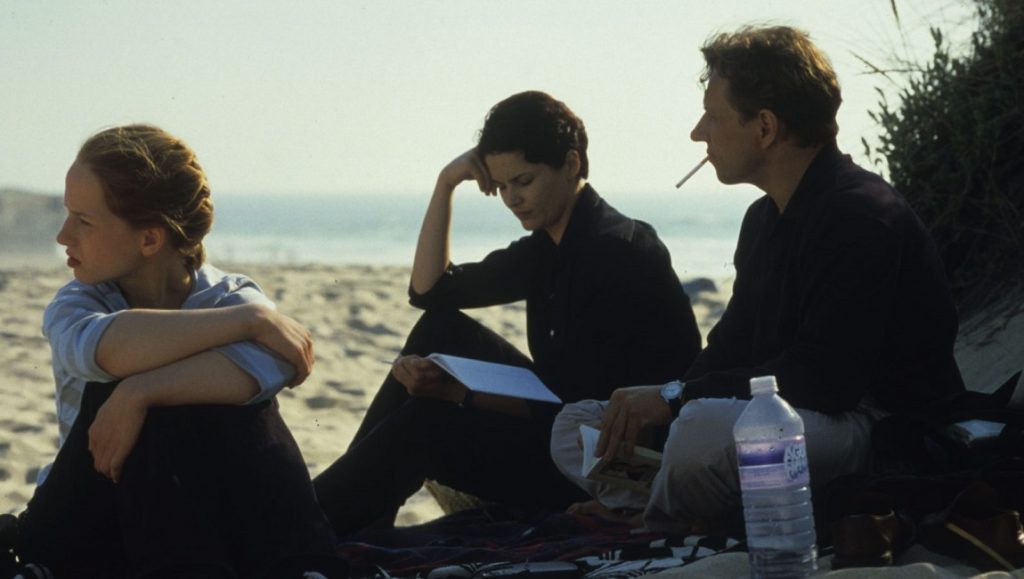 The State I Am In | Christian Petzold Three people sit on a beach. Woman reads, man smokes. Beach scene.
