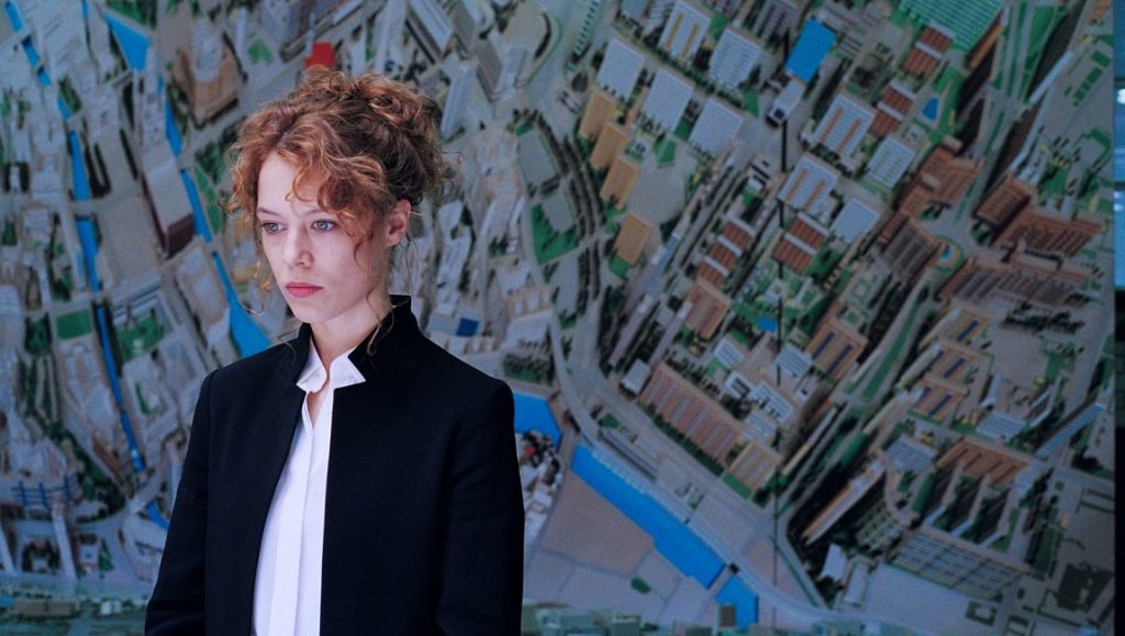 Paula Beer in Undine. Woman in suit stands before city model. German cinema.