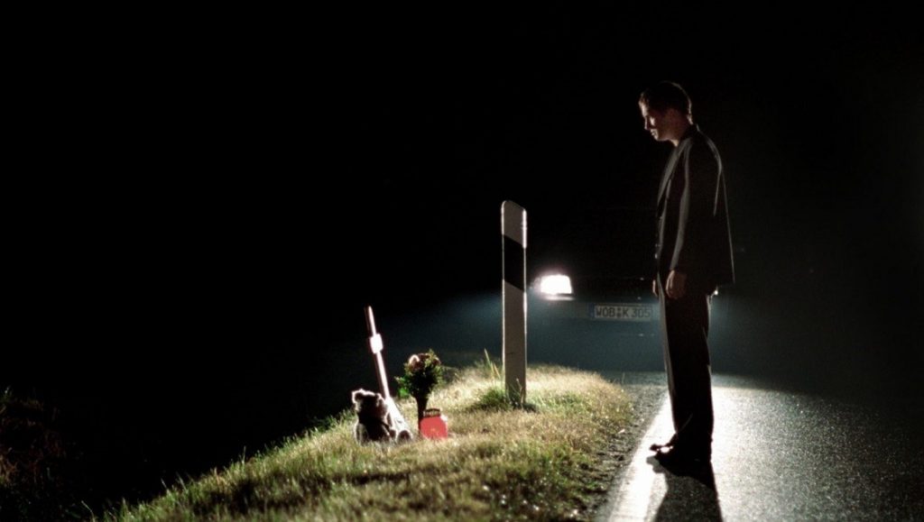 Wolfsburg | Christian Petzold Wolfsburg film still. Man stands on a dark road near a roadside memorial with a car's headlights in the background.