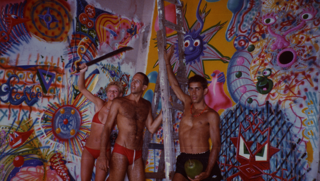 Kenny Scharf art still. Three figures pose in front of a colorful, surreal mural with cartoonish elements. June 2021.