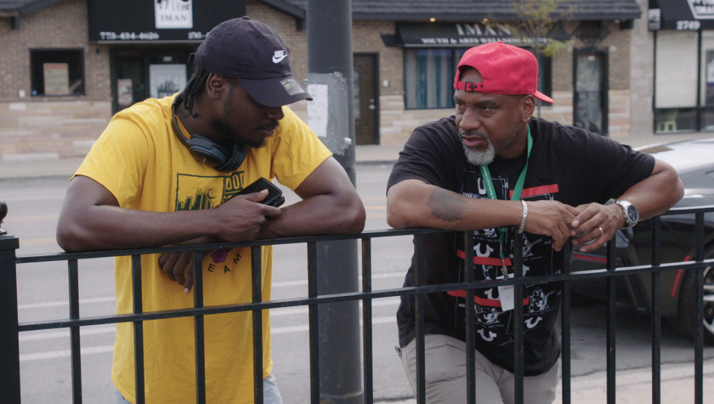 Tribeca Film Festival 2021 — Dispatch 3: All These Sons, Agnes, Shapeless Two men in hats lean on a fence, conversing outdoors. Urban scene with buildings in the background.