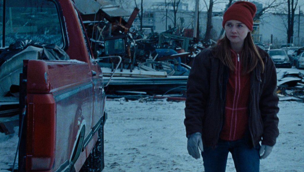 Holler | Nicole Riegel Holler movie still: A young woman in a red beanie stands near a red pickup truck in a snowy junkyard.
