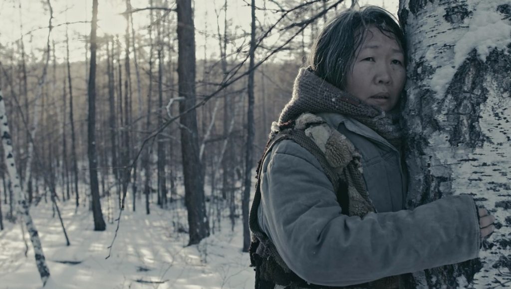 Film still: Woman in winter coat clings to birch tree in snowy forest. Scarecrow movie scene.