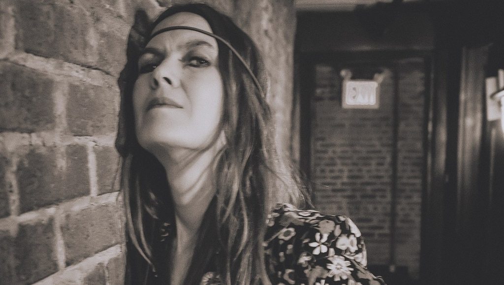 Juliana Hatfield | Blood Monochrome portrait of musician Juliana Hatfield against a brick wall, wearing a floral dress and headband.