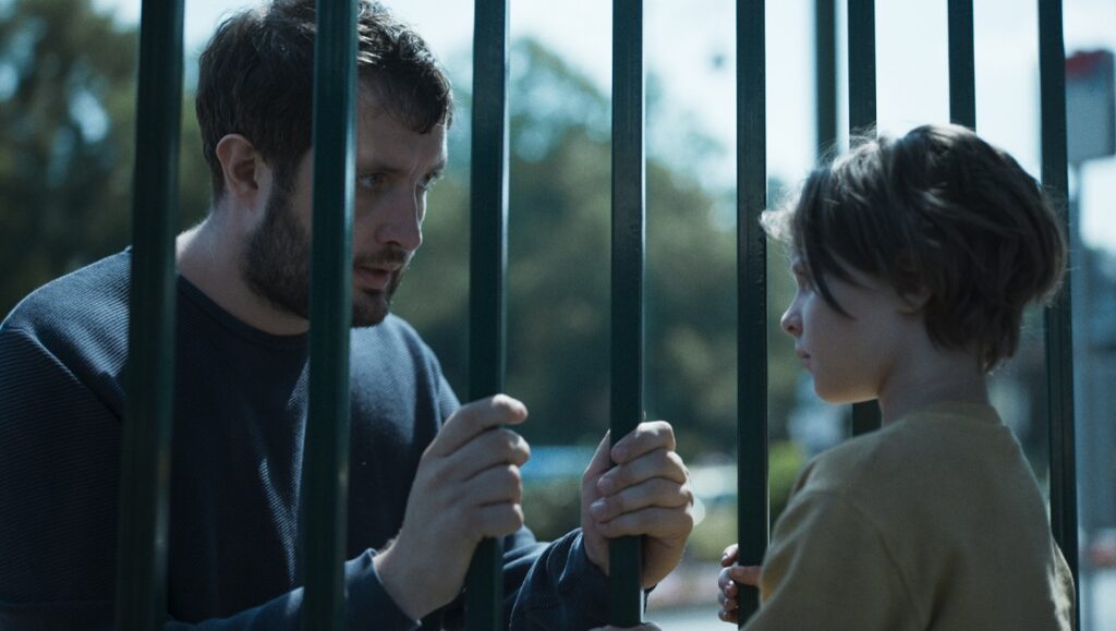 Playground film still: Father and child separated by playground gate. Emotional scene.