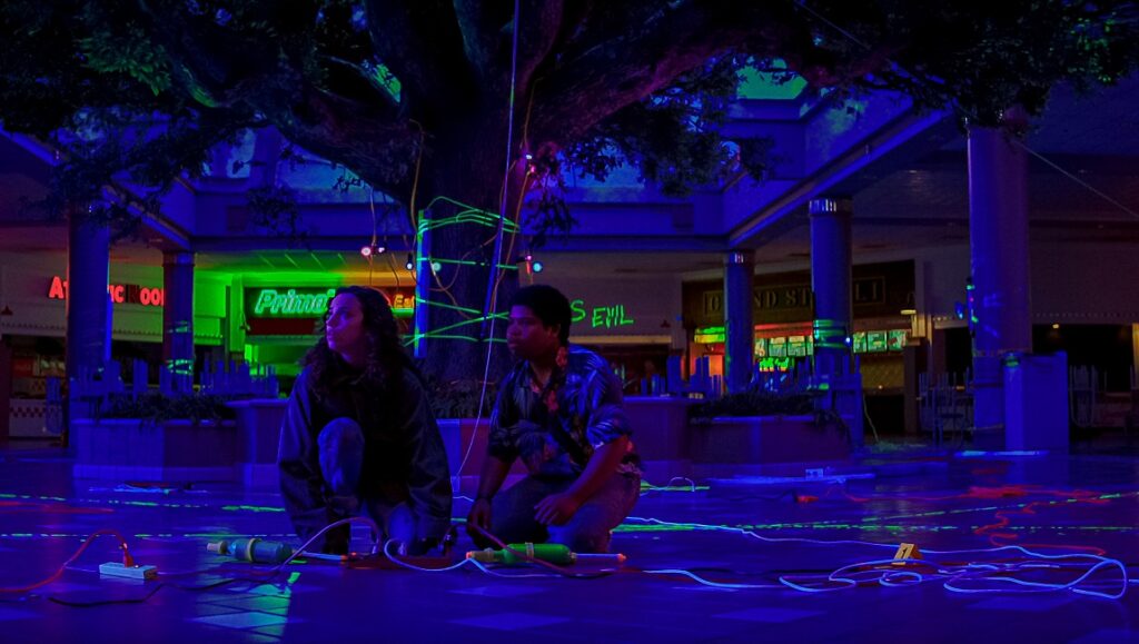 Fear Street Part 3: 1666 | Leigh Janiak Fear Street Part 3: Scene with two people in a mall, illuminated by blue and green lights.