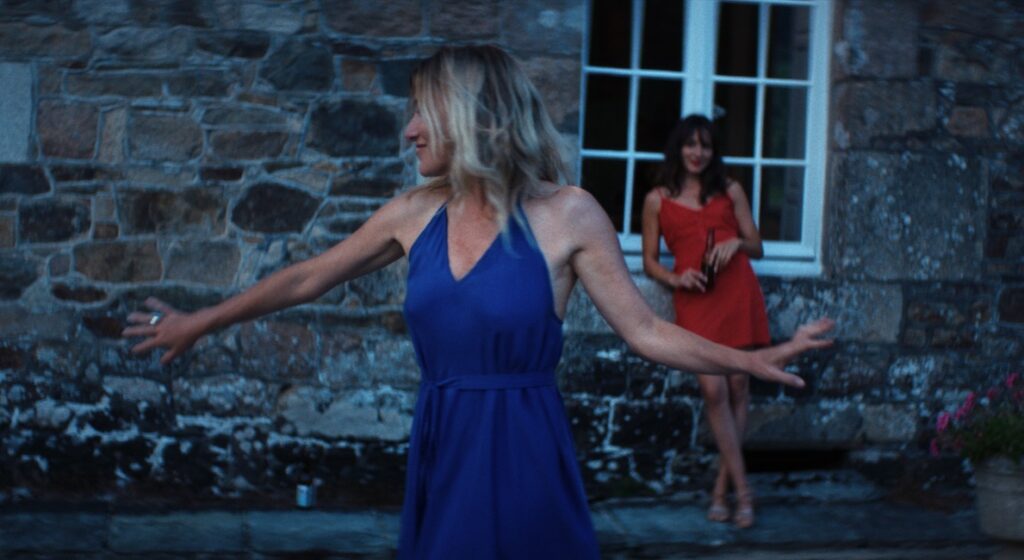 Two women in dresses. One in blue with arms outstretched, another in red by a window. Anais in Love film still.
