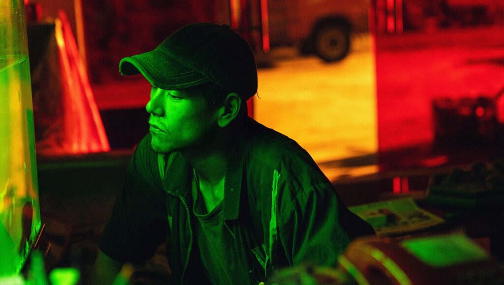 Are You Lonely film still, Cannes Film Festival. Man in cap lit with green and red light. Moneyboys, Medusa, A Radiant Girl.