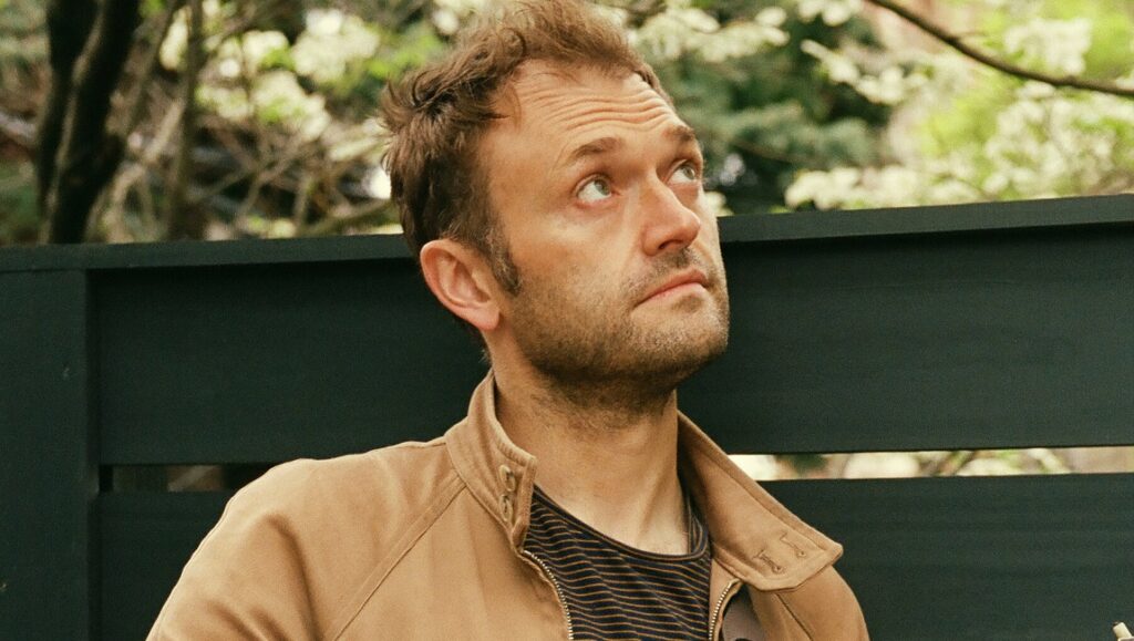Chris Thile, featured on Faye Webster's I Know I'm Funny haha album. Man looking up, wearing a brown jacket.