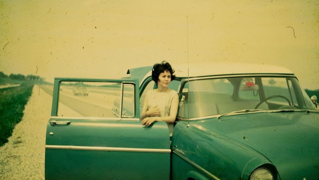 Vintage photo: Woman stands by a classic green car with the door open on a roadside. Travel aesthetic.
