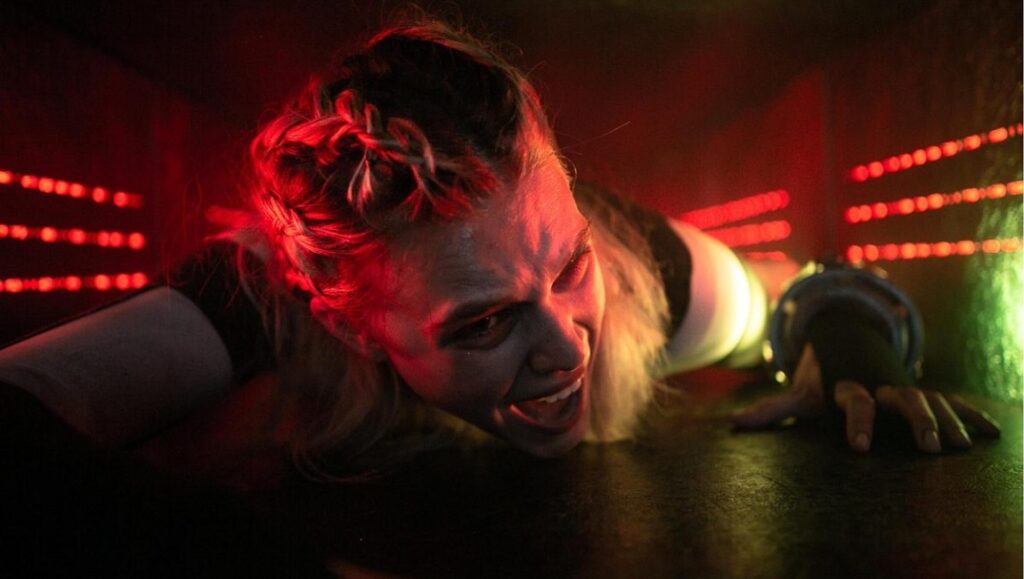 Meander film still. Woman crawls in a claustrophobic, red-lit tunnel. Suspenseful thriller scene.