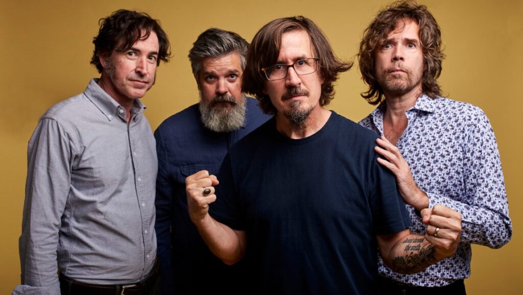 The Mountain Goats band portrait. Four members pose against a yellow backdrop. Indie rock group.