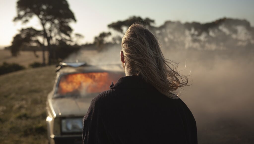 Nitram — Justin Kurzel Nitram film still. Woman with long hair faces away from a burning car in a field. Justin Kurzel film.