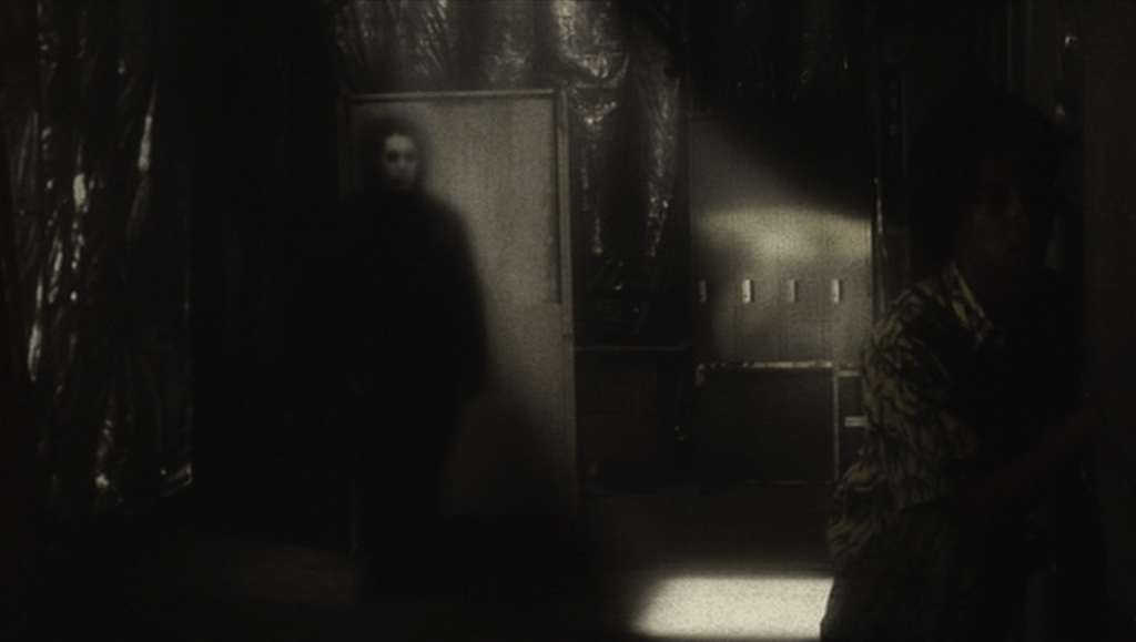 Scene from Kiyoshi Kurosawa's horror film, *Pulse*. Dark figure in a dimly lit corridor.